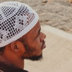 Home Side profile of an adult man wearing a white crocheted cap, sitting outdoors in thought.